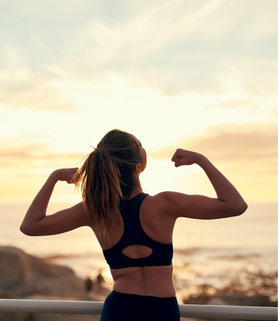 A woman in workout attire with her back to the camera flexes her muscles against a beautiful sunrise