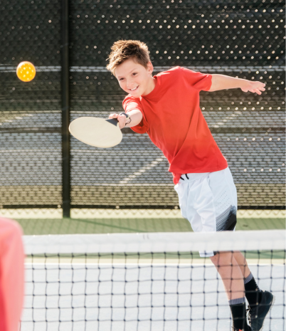 A young boy wearing a red t-shirt plays pickleball with a friend.