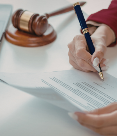A female hand signs a document. On the desk are an open laptop, and a gavel.